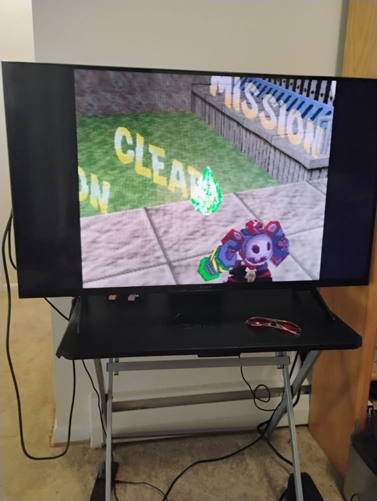 Scene from The Misadventures of Tron Bonne for the Sony PlayStation (PS1 or PSX) on a flat screen. A dark red robot looks at a green gem, as a nondiegetic "Mission Clear" disappears from the screen.