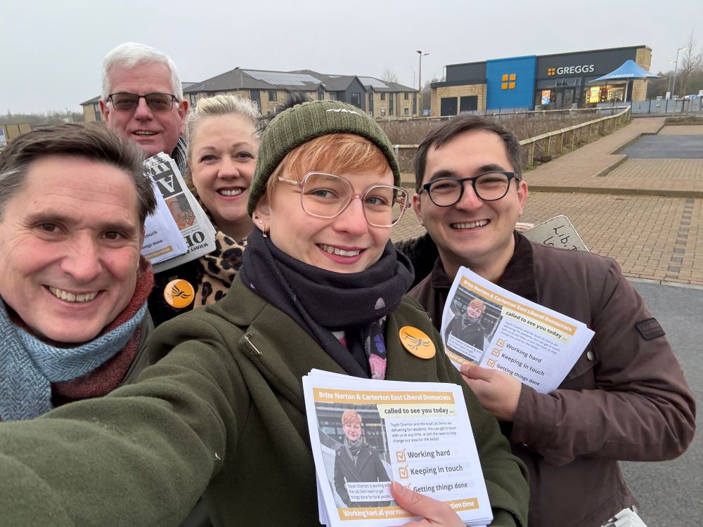 A group of five smiling Liberal Democrat campaigners holding leaflets while canvassing, with a Greggs store visible in the background.
