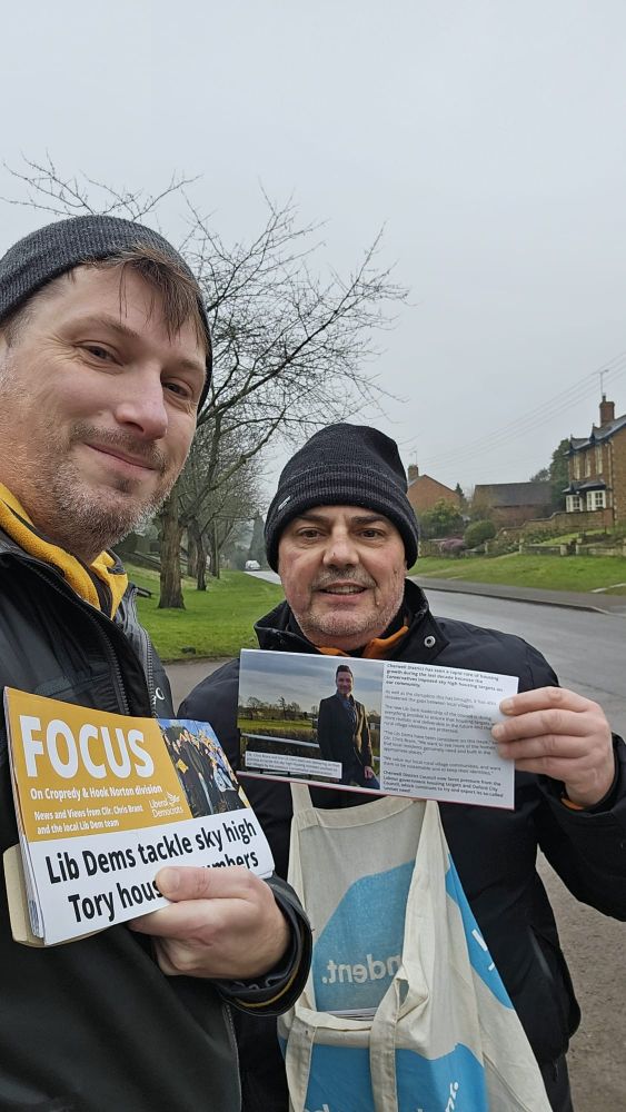 Two men in winter hats holding Liberal Democrat FOCUS newsletters, standing on a residential street with trees and houses in the background
