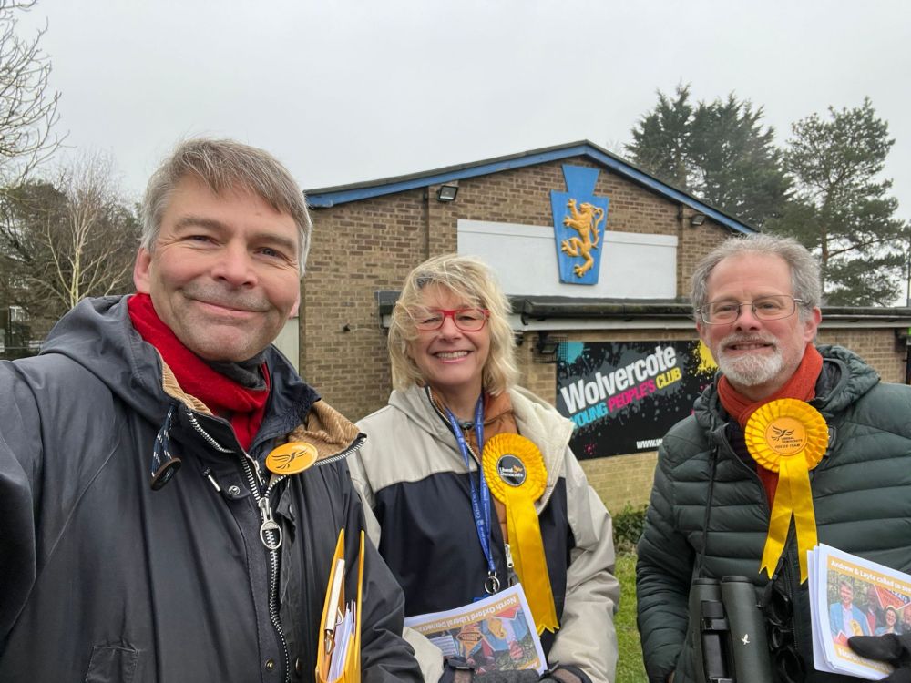 Three Liberal Democrat campaigners wearing yellow rosettes and holding leaflets, standing outside Wolvercote Young People’s Club.