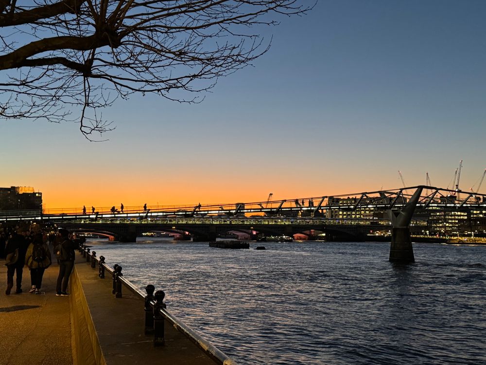 A view of sunset behind the millennium bridge London - stretching over a relatively calm Thames. The orange glow is offset by blue of the water and upper levels of skyline. Some pedestrians can be seen both on the sidewalk and the bridge. 