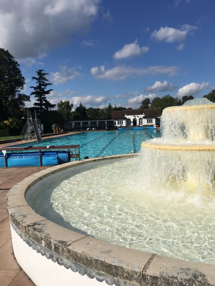 A historic 50m outdoor pool, with a large fountain in the foreground, on a sunny summer day.