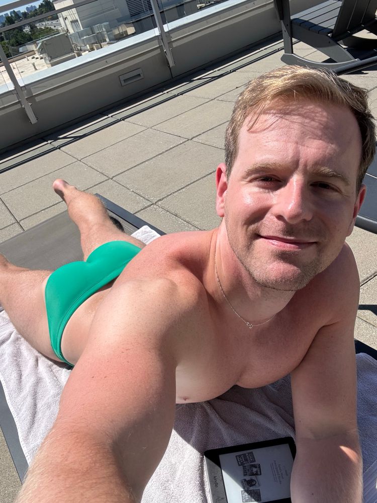 Guy taking selfie while laying face down on lounge chair. It’s sunny and he’s wearing a green speedo 