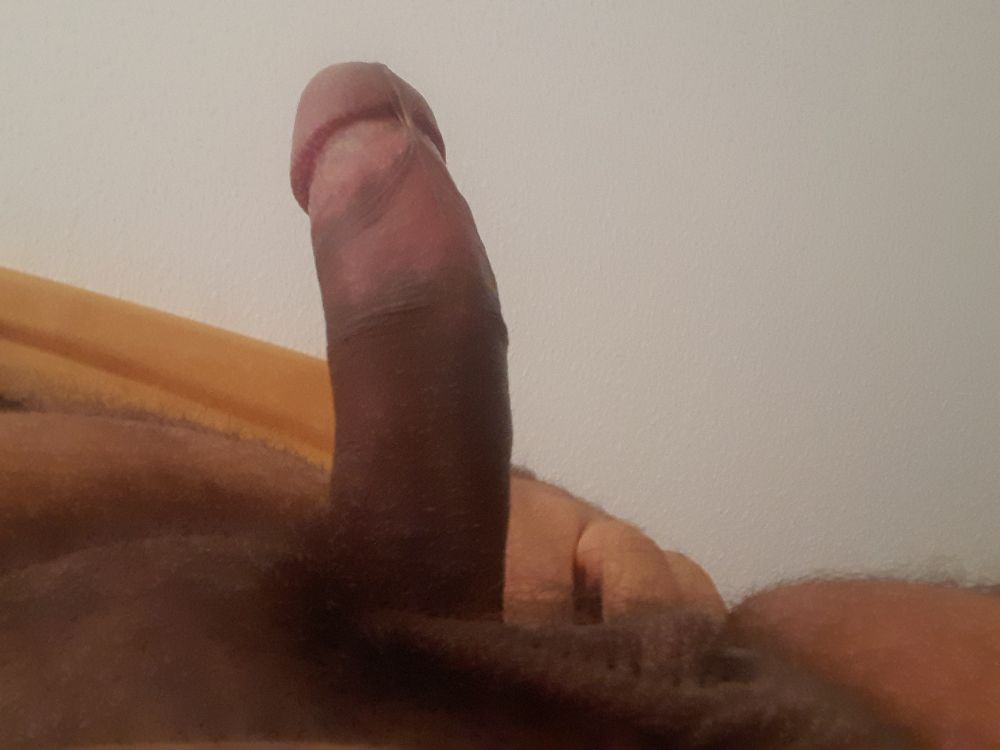 An erect, brown, uncut penis, foreskin fully retracted. You can see some sack, as well as the owner's hairy fupa and the underside of his belly. In the background the headboard of a bed and a white wall are visible 