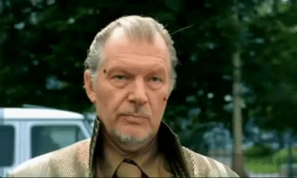 a man with gray hair and a beard is standing in front of a white van and looking at the camera .