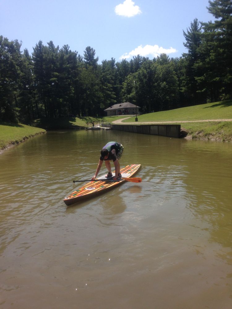 Person in a striped shirt leaning over as they balance on a paddle board in a pond. Trees and a small cabin in the background 