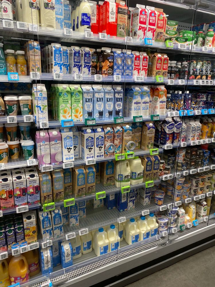 Small shelf showing the refrigerated milk section of a small Dutch supermarket. At least 25!different types, sizes, and brands of milk and milk-like drinks are visible. 