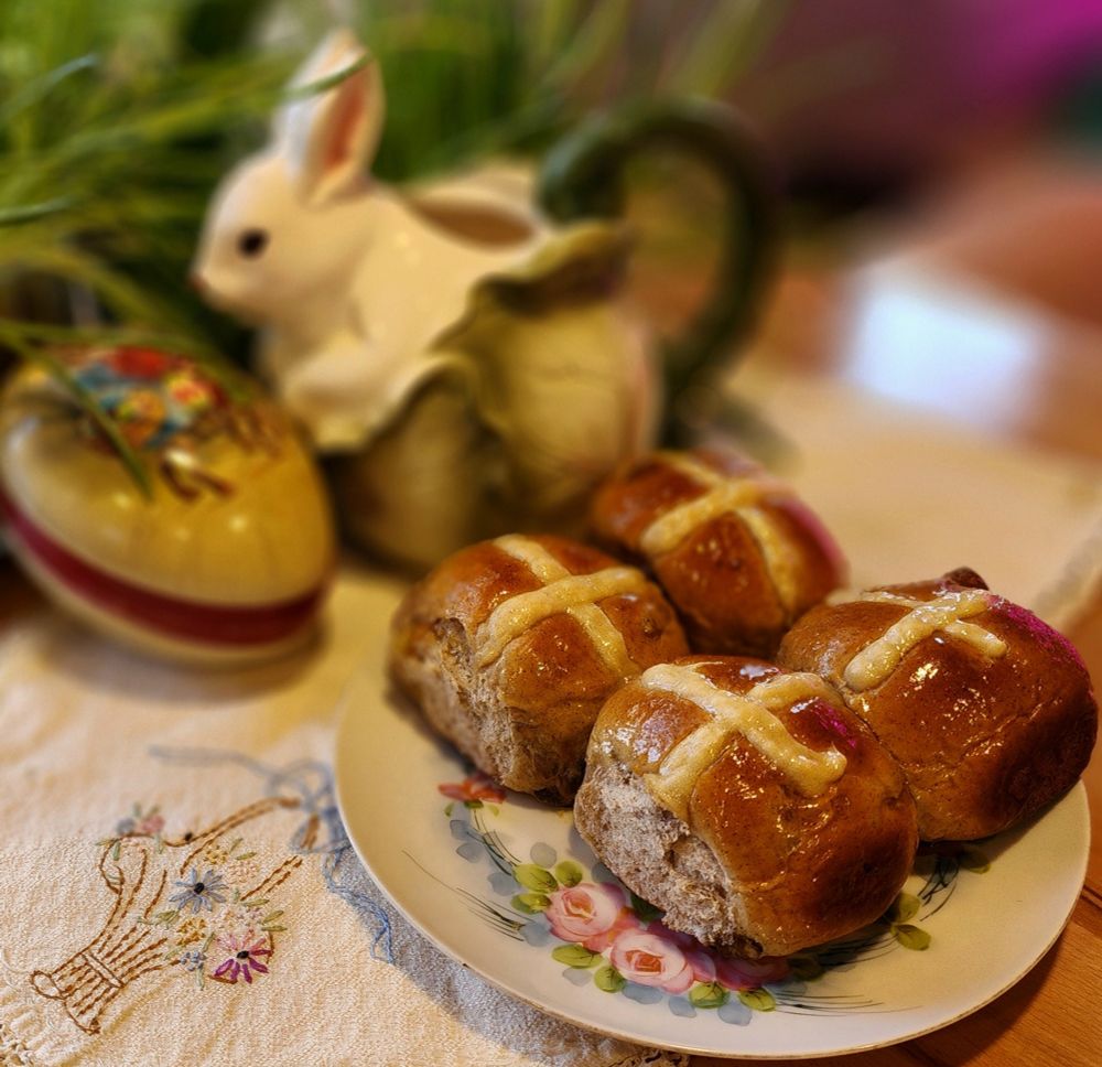 4 Hot Crossed Buns on a plate, sitting on an embroidered cloth. An Easter egg, a rabbit-shaped pitcher, and some greenery are in the background. 