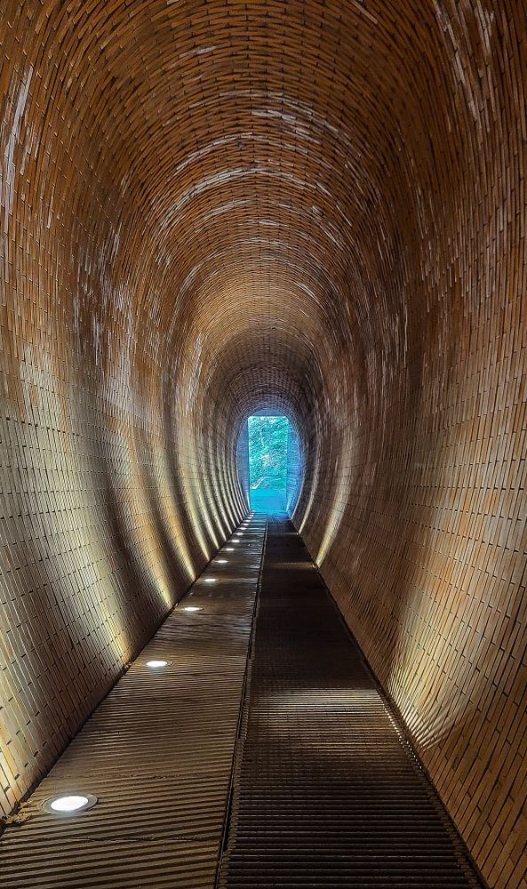 Long narrow oval shaped tunnel lighted by round yellowish reflectors built into the floor which project the soft light beams onto the walls and ceiling. Daylight is seen in the distance at the end of the tunnel. 