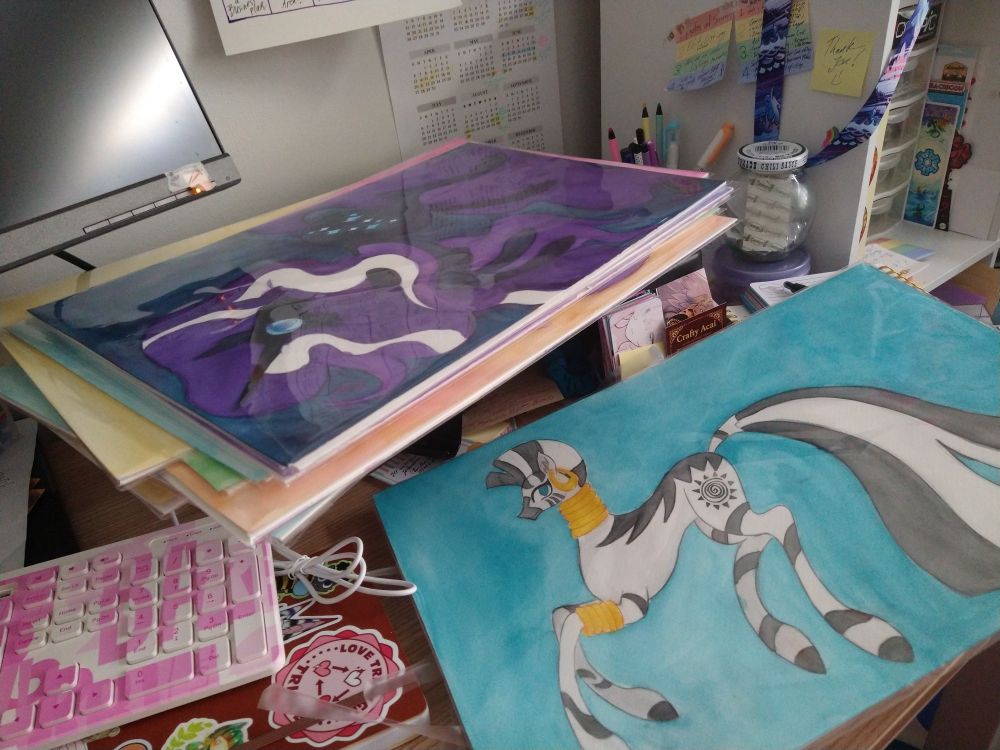 Photo of a messy desk, mostly covered by large watercolor paintings of my little pony characters. (Nightmare rarity, and zecora specifically)
