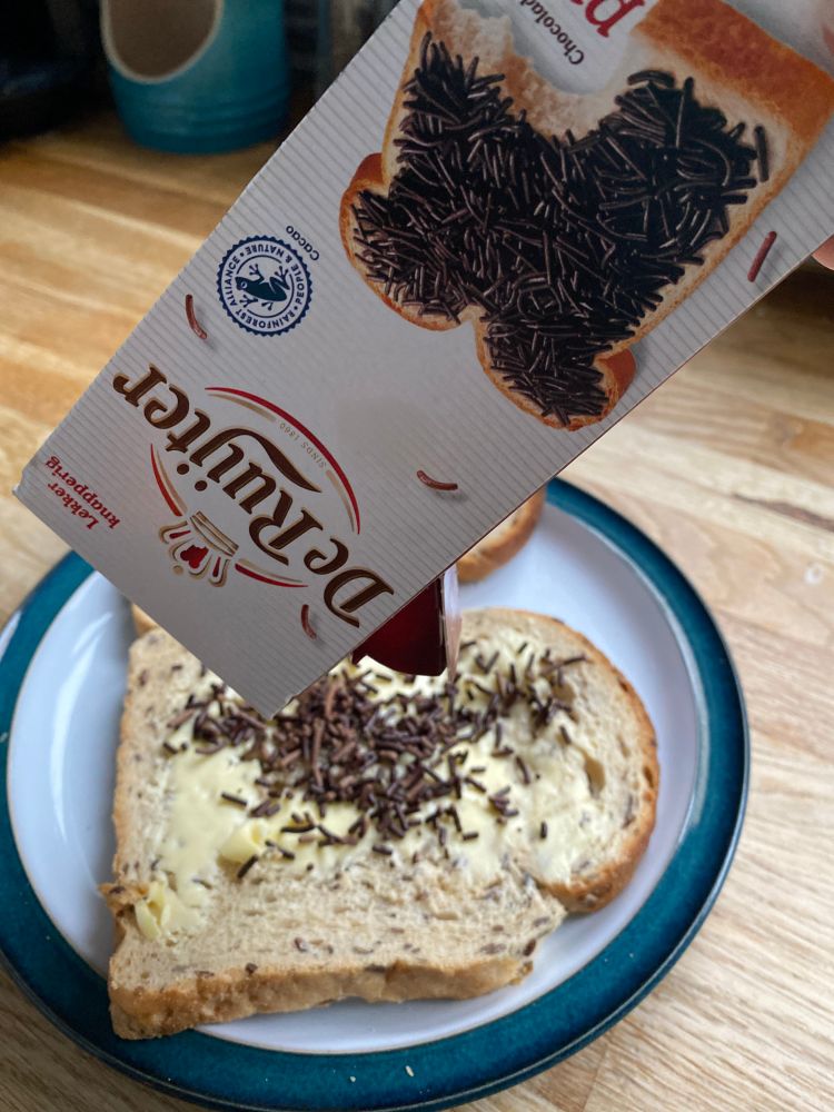 An empty box of Dutch chocolate sprinkles (hagel slag) held over some buttered bread. Only a few sprinkles made it onto the bread before they ran out. 