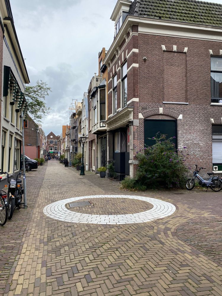 A small dutch street with brick block paving and a T junction. There is a white circle outline painted in the junction like a roundabout but it goes nearly to the edge of the carriageway. 
