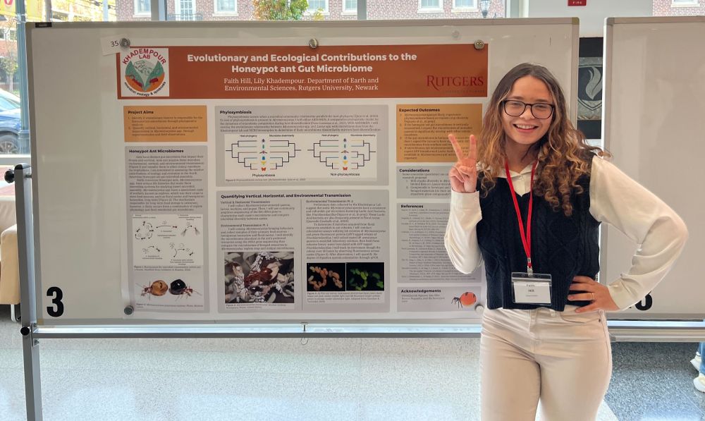 A woman stands, smiling, making a peace sign, in front of a poster titled, "Evolutionary and Ecological Contributions ot the Honeypot ant Gut Microbiome"