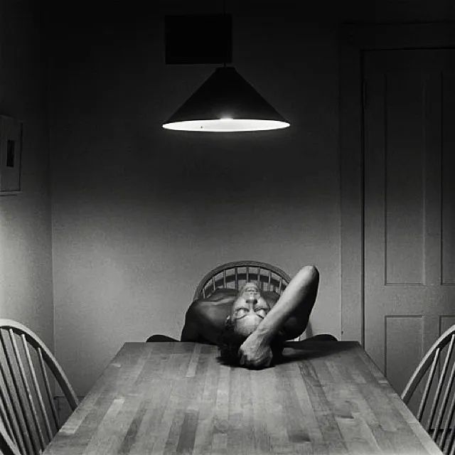 A Black woman leans her back against a wooden kitchen table. Her right hand holds her hair with her arm bent. 