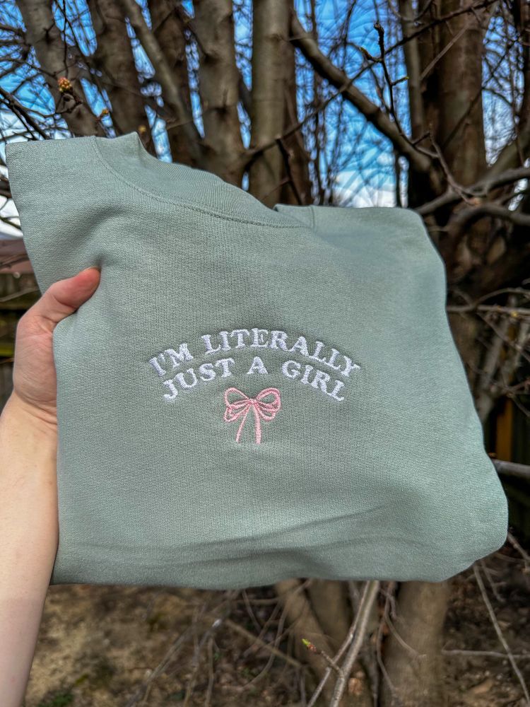 A sage green embroidered crewneck reading “I’m literally just a girl” in white text with a pink bow 
