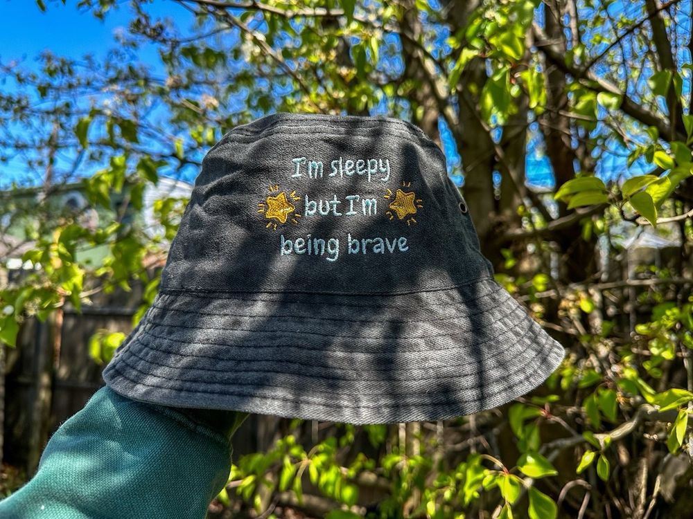 An embroidered bucket hat photographed outside which reads “I’m sleepy but I’m being brave” 