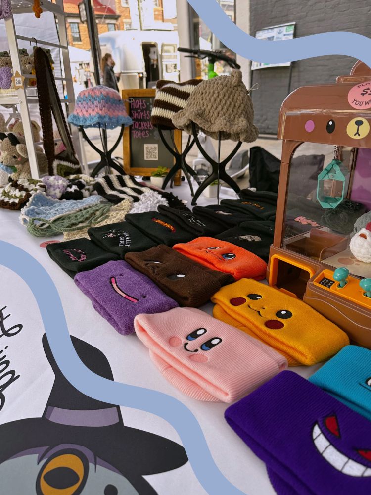A collection of embroidered and crocheted beanies and bucket hats at an art booth at a market event- Pokemon, Kirby, funny sayings 