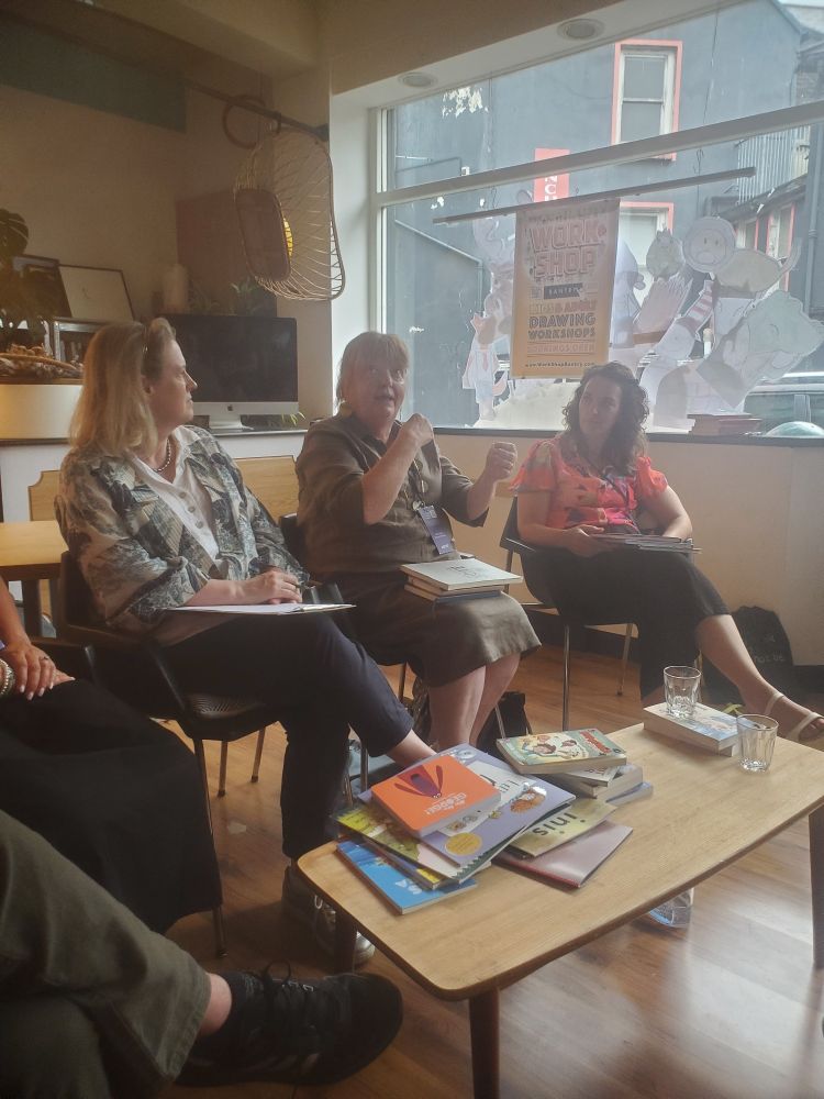 Patricia Forde speaks to an audience in the Work Shop. She has red hair and wears a brown dress. Sarah Webb is to her right, and Leona Forde is to her left. In front of her, a table holds loads of children's books.