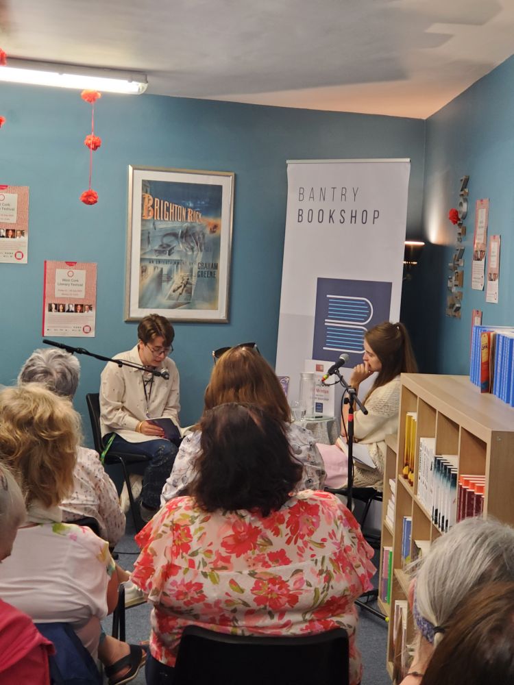 Kel Menton reads from his novel A FIX OF LIGHT into a microphone to an audience at Bantry Bookstore. Molly Twomey is to their left. Kel wears a white shirt and glasses and has short, fair hair.