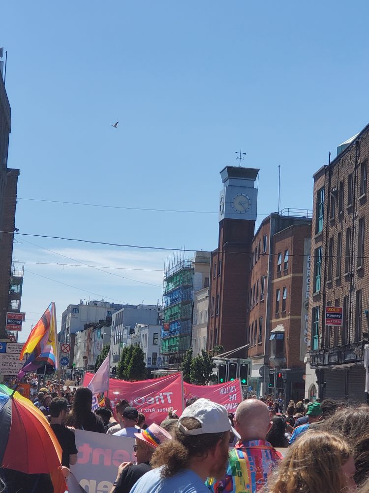 The Limerick Pride parade on O'Connell Street under a clear blue sky, with one bird in flight.