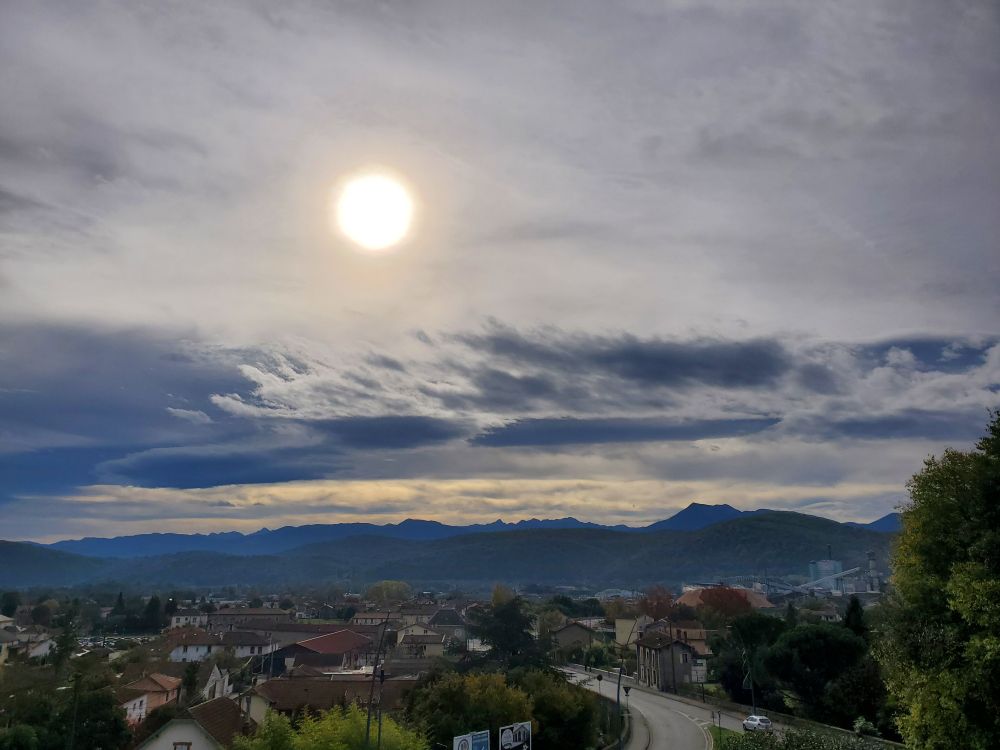 Photo de soleil dans le ciel brumeux, avec la ville en contre bas, et la ligne des Pyrénées 
