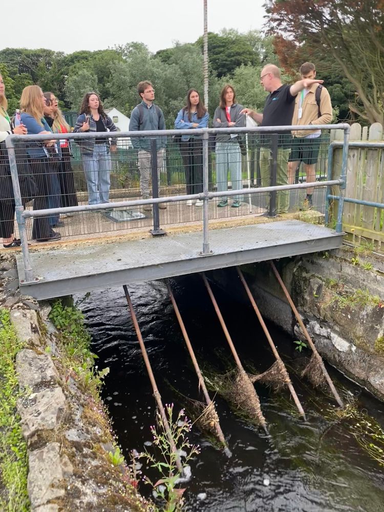 A man explains how salmon are caught and counted on a bridge. A group of about 8 scientists (most women) are listening.