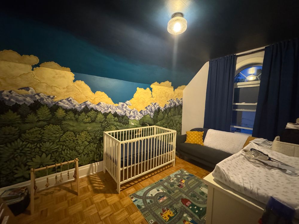 A kids room with a mural on the wall, crib in the middle, and couch against the window 
