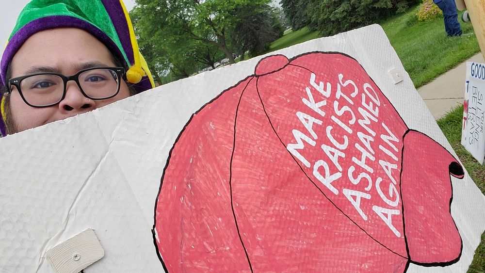 Person wearing a jester hat holding a sign that says "Make Racists Ashamed Again“