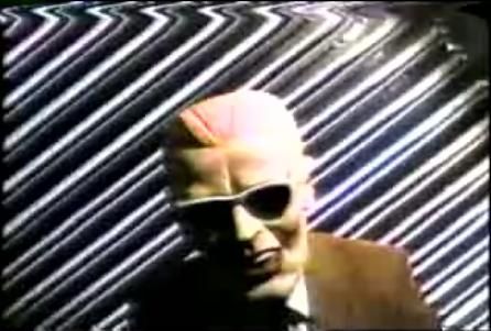 A screenshot from the infamous Max Headroom Incident, featuring an unknown TV hijacker in a rubber Max Headroom mask in front of a corrugated metal background. 