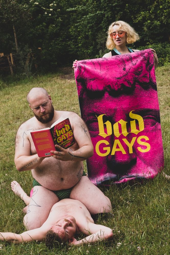 three people in a meadow. One has chin-length blonde hair and is wearing sunglasses and what looks like some kind of halter neck garment. It is hard to tell what they are wearing because they are holding a BAD GAYS beach towel in front of them. The towel has a close up image of a Greco-Roman statue colored pink on it, with the words BAD GAYS imposed in yellow. The other two people are in front of the person holding the towel. The skinnier one lies on the ground, at least shirtless, possibly naked. The third person, clad only in what looks like a Speedo, straddles the person lying down, while reading the BAD GAYS book, with an identical cover to the beach towel. 