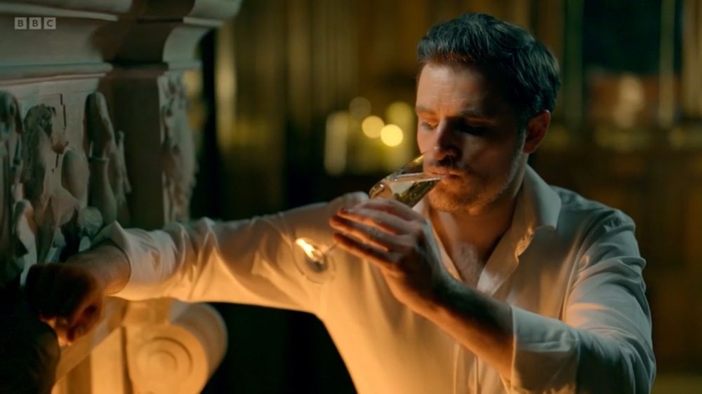 Alexander from The Traitors looking fit, leaning against a fireplace drinking champagne.