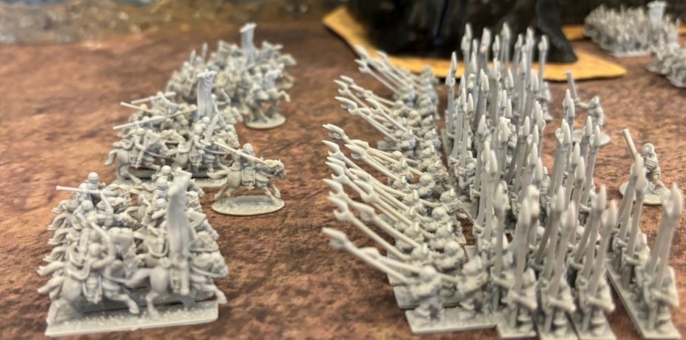 Numerous 10mm scale gray miniatures are arranged in formations on a textured surface, depicting armies of orcs and humans for tabletop gaming.