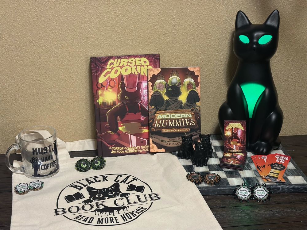 A smattering of goodies from Cat Eye Press including Cursed Cooking and Modern Mummies anthologies; various earrings; a cute tote bag, and more! All while a glowing eyes cat looks on. 