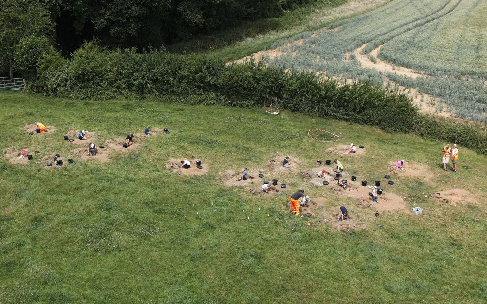 Overhead shot of the excavations. Around 20 children dig in individual test pits, supported by archaeologists, teachers and helpers.