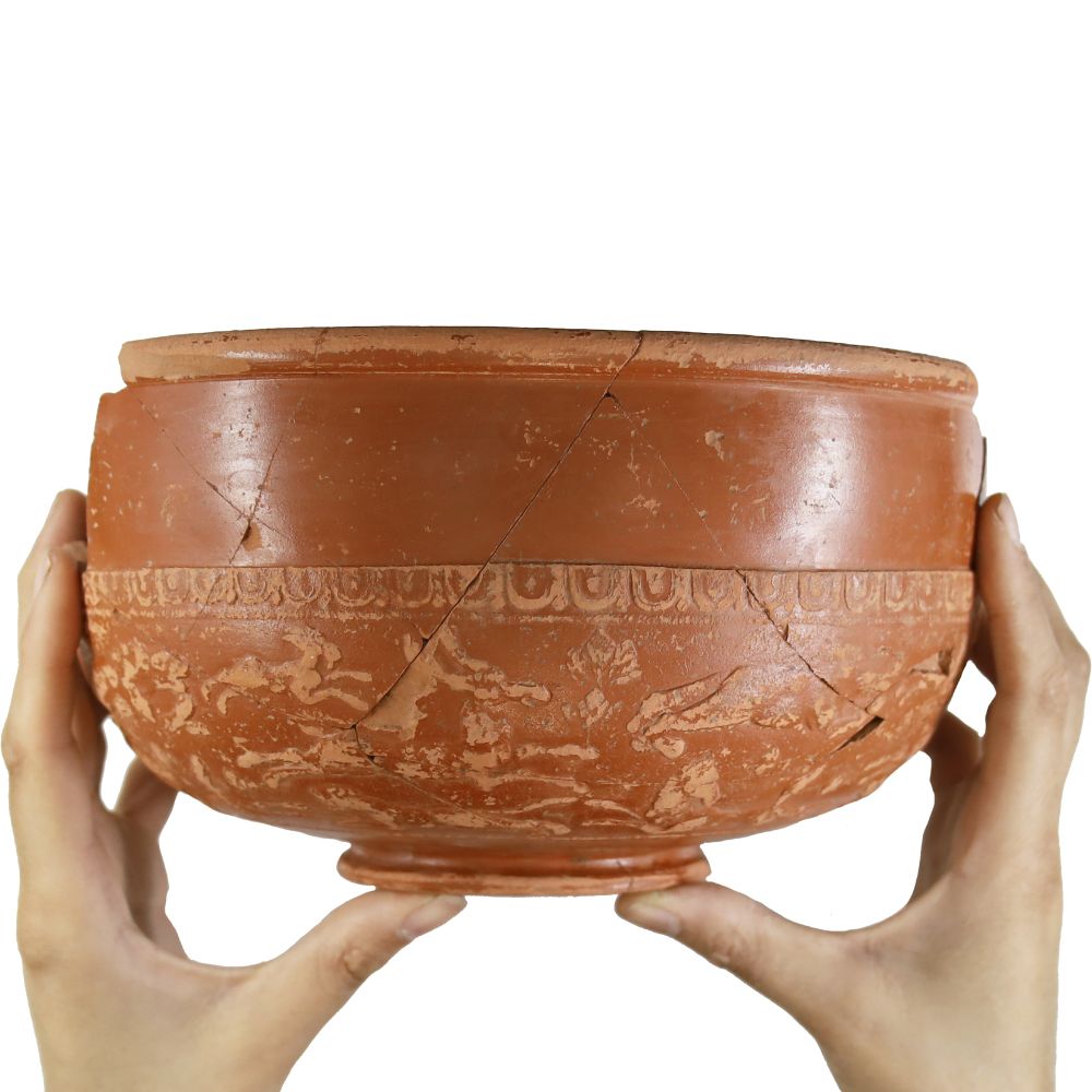 Two hands hold up a red ceramic bowl with a pattern around the edge of hares running. This type of pottery is known as Samian.