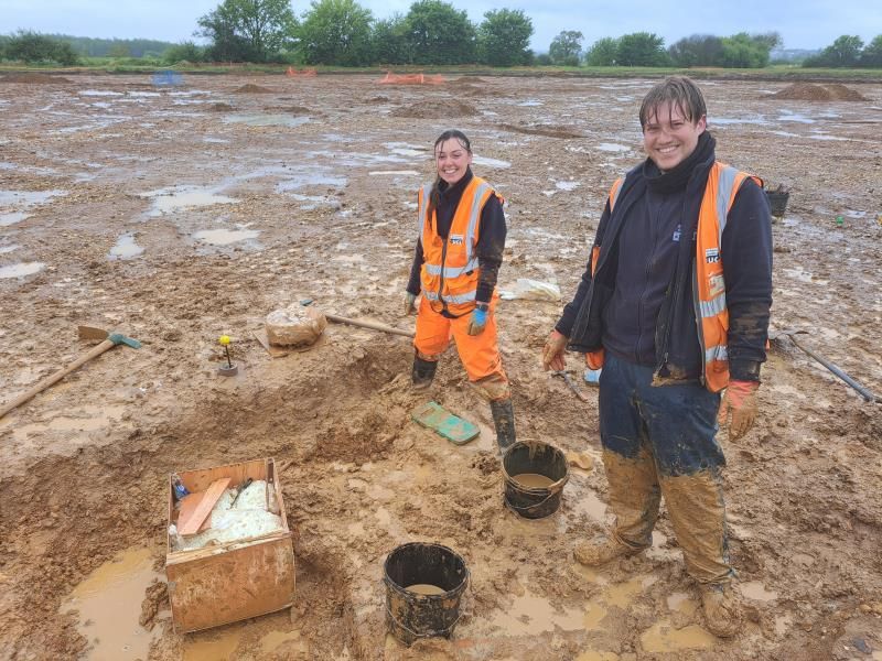 Two very damp and muddy archaeologists grin at the camera. They are standing with tools and buckets on an archaeological site that has clearly just been hit with a downpour. Muddy puddles surround the area where they are working.