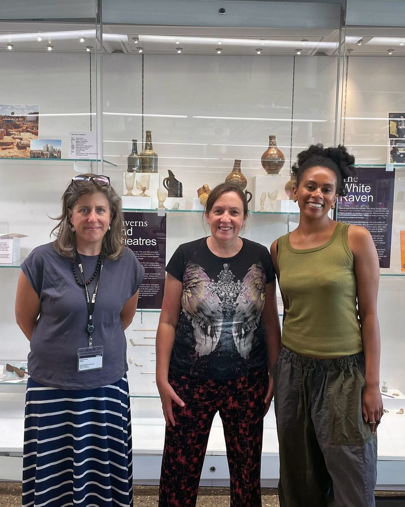 Three women stand smiling in front of an exhibition case. Behind them you can see archaeological wine glasses, bottles and ceramic jars, alongside labels in a glass case.