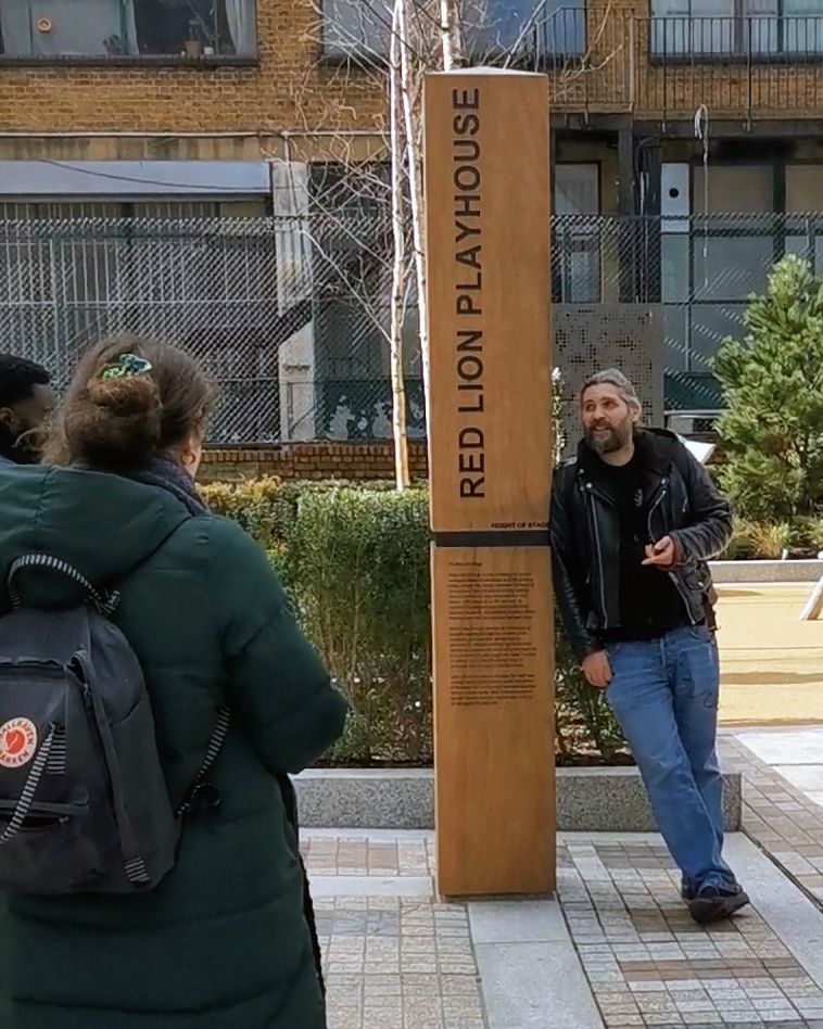 A man chats to people while leaning against a tall wooden post that reads “Red Lion Playhouse”
