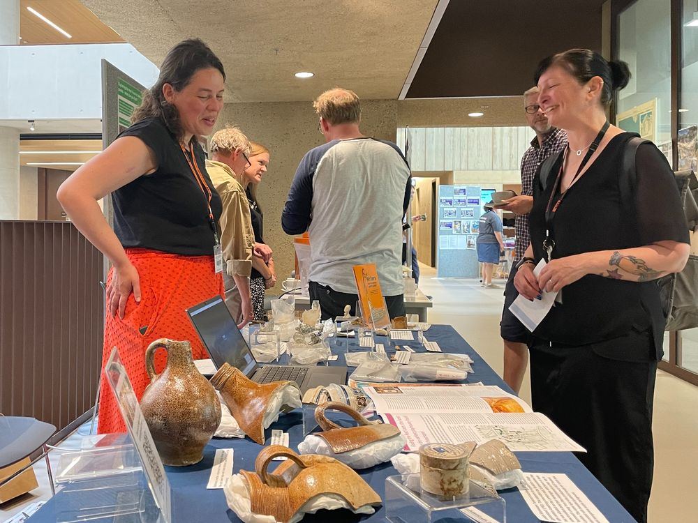 A woman standing behind a table full of archaeological artefacts talks to people visiting the stand