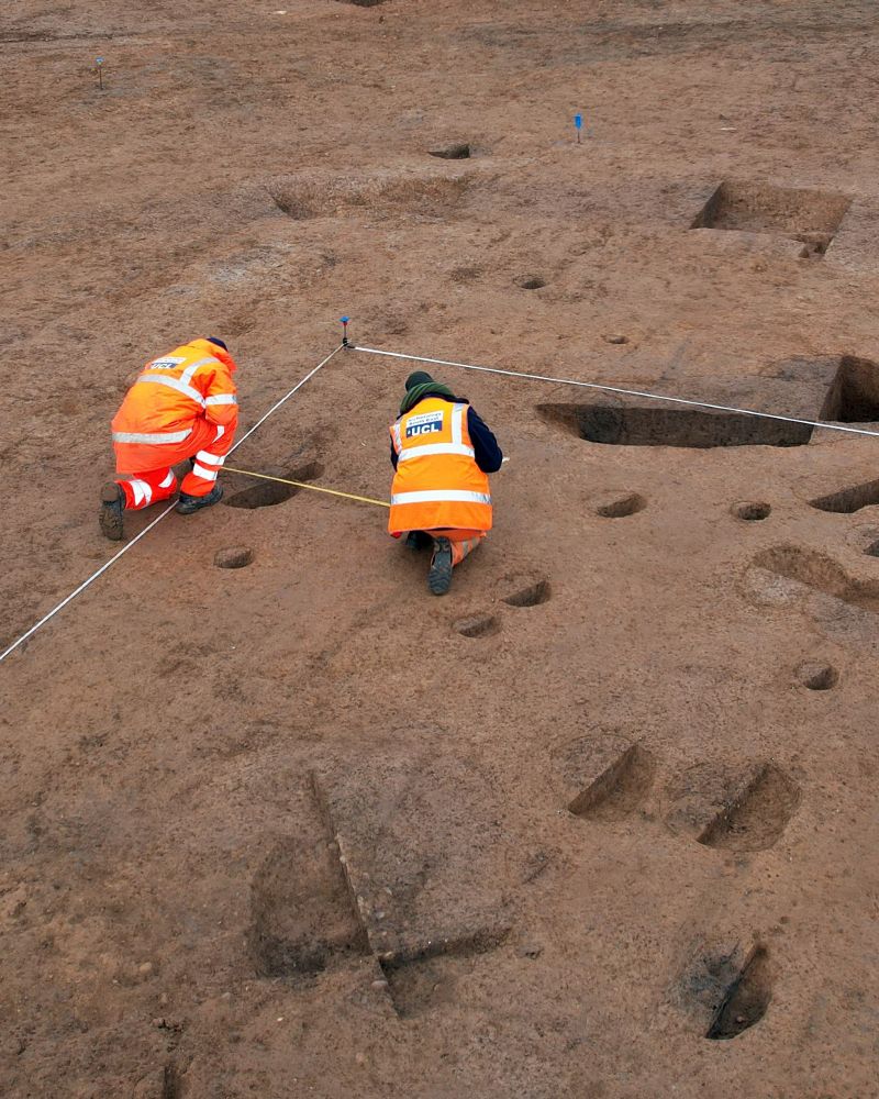 Two archaeologists seen from behind working on an archaeological site, surrounded by lots of holes indicating excavated features.