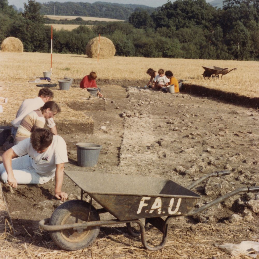 A slightly sepia toned photo of an excavation from the 1980s. A wide, shallow trench has been dug in a field with large hay bales, and multiple people sit in lines in the trench working with trowels. In the foreground a wheelbarrow bears the letters FAU.