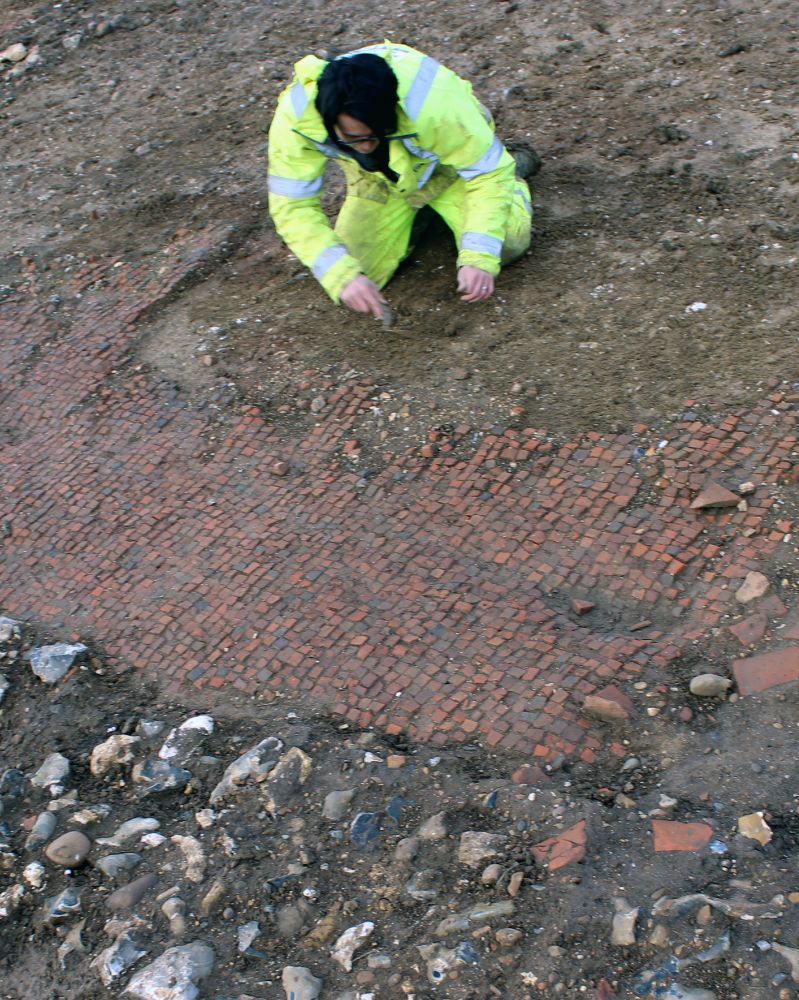 An archaeologist carefully excavates an archaeological floor of many small square tiles.