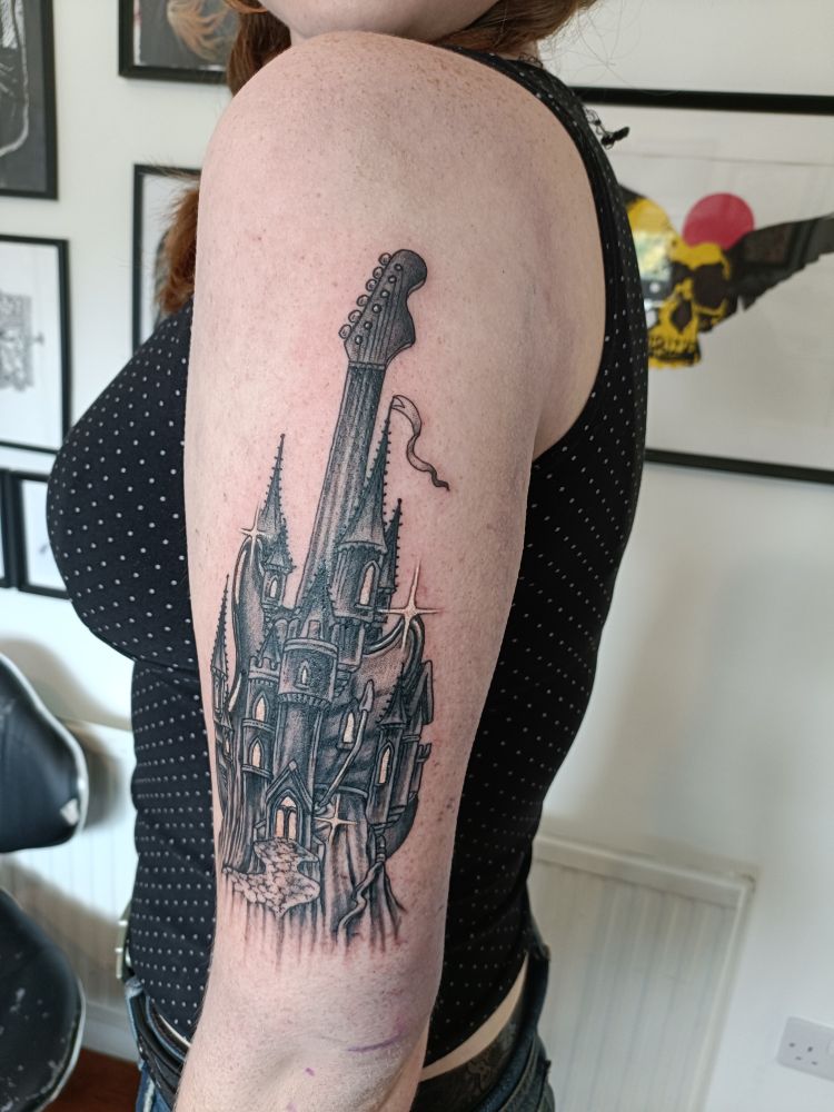 My daughter's freshly-tattooed upper arm. The large tattoo, stretching from shoulder to elbow, depicts a gothic-style castle with a guitar neck emerging from the top as if it were a turret. The image is based on the cover art for Ritchie Blackmore's Rainbow, but has been reinterpreted in blackwork by the Irish tattooist Joseph Deegan. 