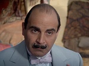 A photograph of Hercule Poirot taken from slightly above.  He looks up.  He is balding, has brown eyes and a black mustache that is elaborately upturned at the ends. He wears a light blue suit and a bow tie.