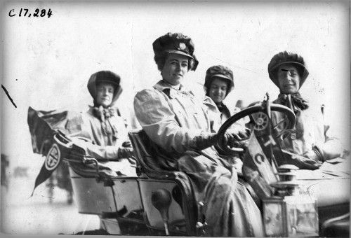 A black-and-white photo of four young women sitting in an open motor car.  They are wearing hats and looking at the camera.  The one in the back left has a pennant hanging next to her.