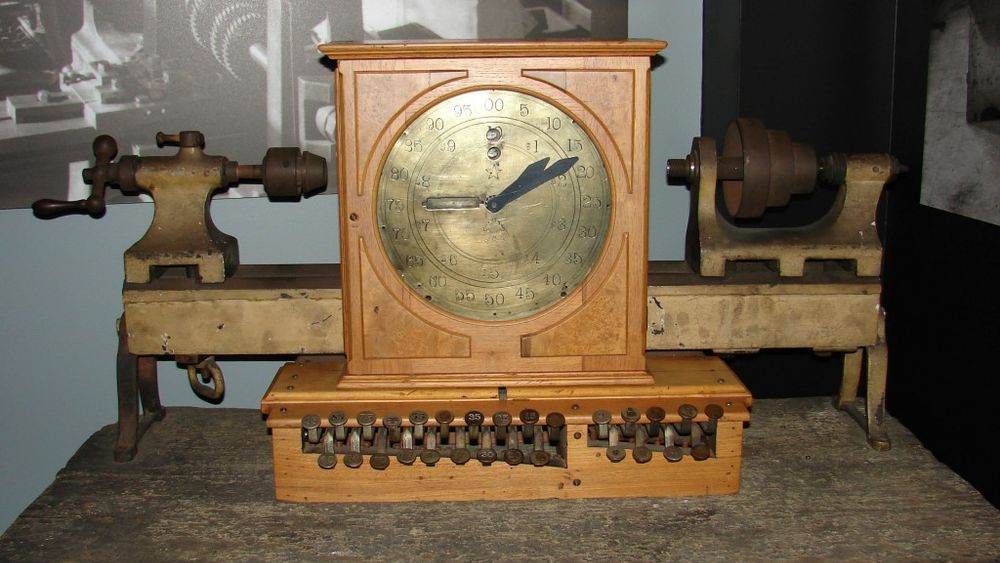 A photograph of the cash register.  It is a wooden box with a gold face like a clock's, with two black hands and one hand that appears to be gold.  The gold hand points to the 9 o'clock position, the smaller black hand points to the 2 o'clock position, and the larger black hand is two minutes past that.  The clock-like box sits on top of a rectangular box with a row of small levers.  The row has a small gap a third of the way from the right-hand side.