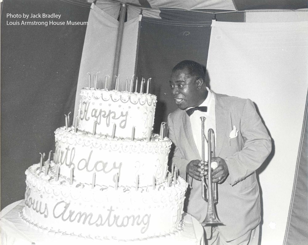 A black-and-white photo of Louis Armstrong standing next to a huge birthday cake.  The cake has 3 tiers.  The top tier says "Happy," the middle tier says "Birthday," and the bottom tier says "Louis Armistrong." there are lots of candles on each tier.  Armstrong wears a light-colored suit with a narrow black bow tie and light-colored pocket square.  He holds his trumpet in both hands and leans towards the cake.