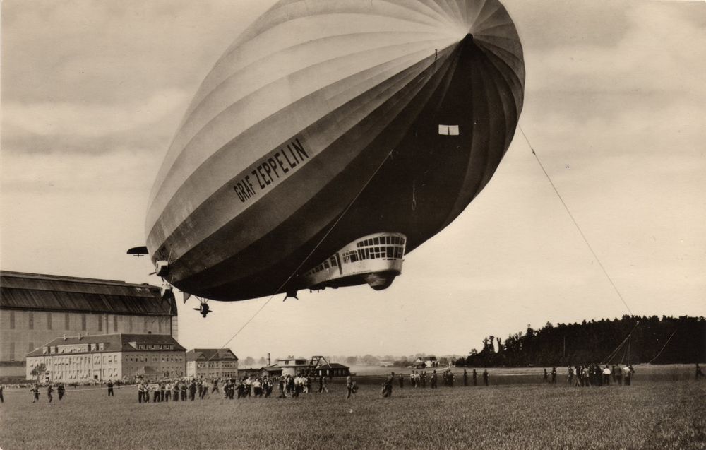 A black-and-white photo of the Graf Zeppelin.  The airship hovers over a crowd of people far below.  There are buildings in the background, and trees very far away