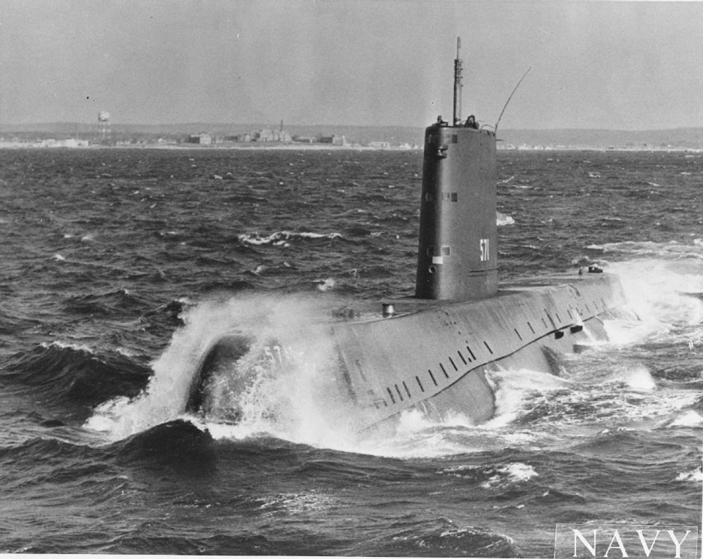 A black-and-white photo of the USS Nautilus, a nuclear submarine, moving through the ocean.  There is a city in the far background, and the word "Navy" in the lower right-hand corner of the picture