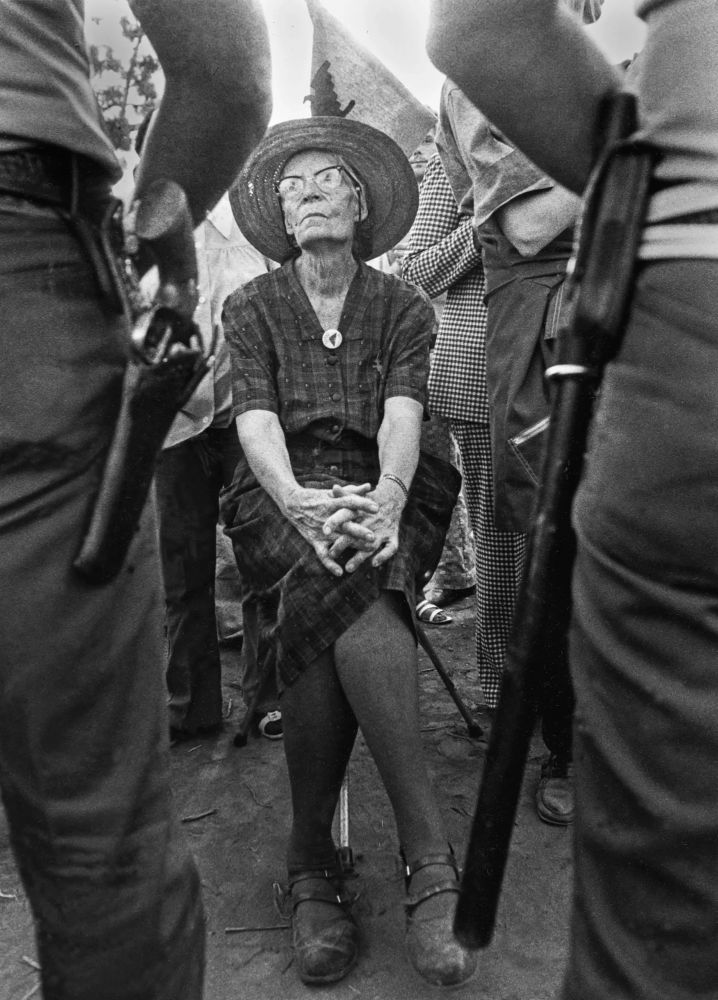 Dorothy Day, elderly, seated in a chair on the frontline of a protest flanked by two armed police in front of her, facing her. She sits calmly hands folded over crossed legs, picture taken from hip high to the police, her in full view 
